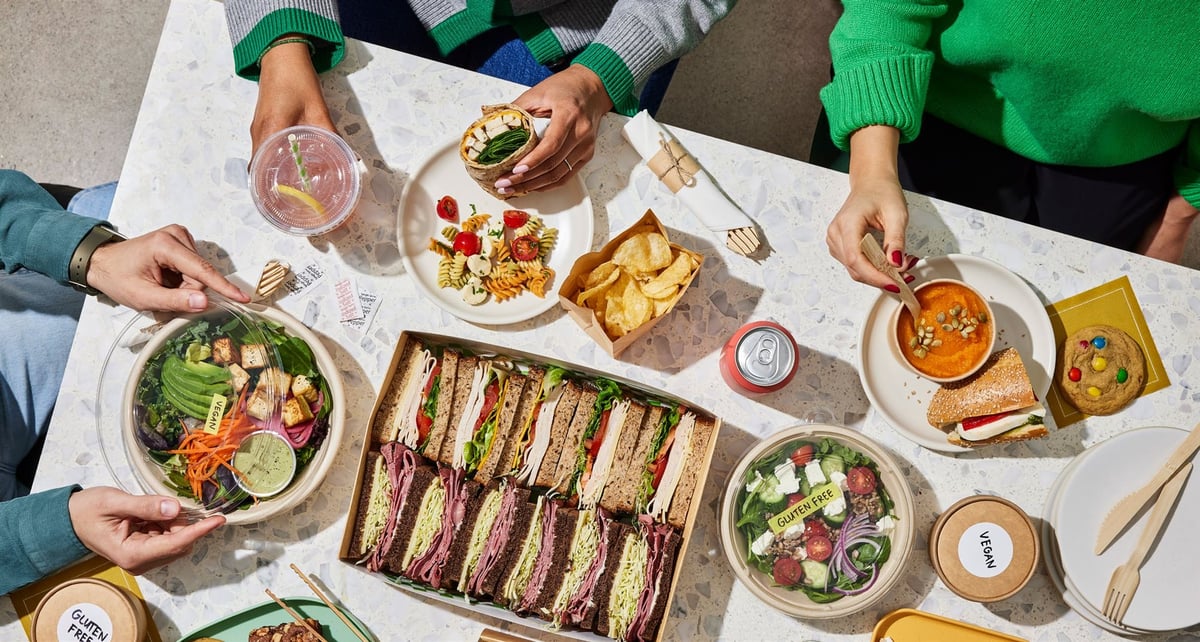 An overhead view of a shared office catering table: a boxed sandwich platter, salad bowls, soup, crisps and a cookie, with three people reaching in