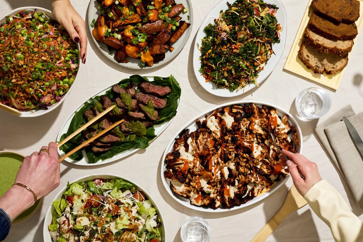 Hands serving from an atis catering spread of salads, roast vegetables, sliced steak and banana bread