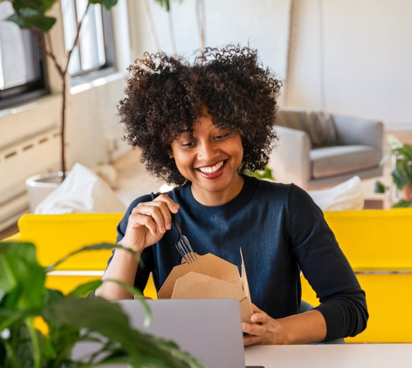 A woman at her desk smiling as she opens a takeaway lunch box, laptop in front of her