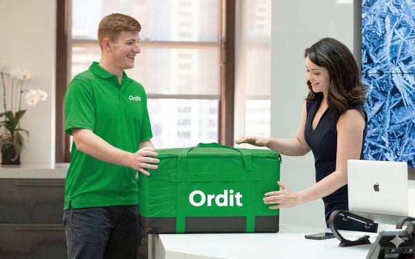 An Ordit courier handing a large insulated Ordit delivery box to someone at an office reception desk
